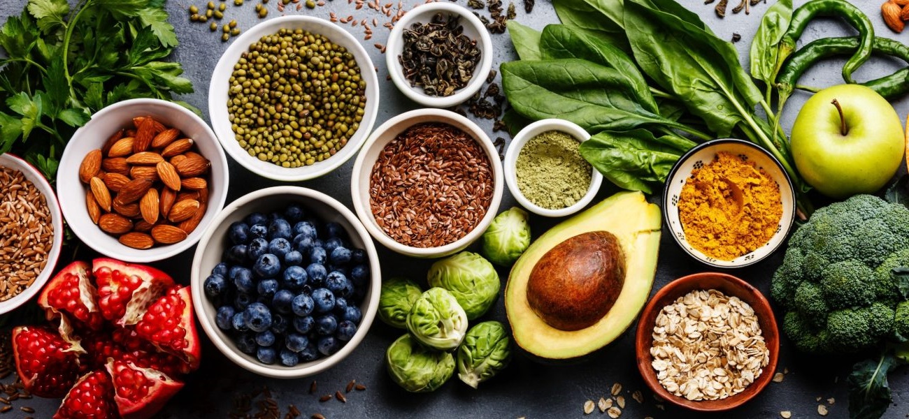 Anti-aging foods for healthy skin, including avocados, blueberries, sweet potatoes, almonds, walnuts, green tea, dark chocolate, tomatoes, and pomegranate seeds, arranged on a wooden table in natural light.